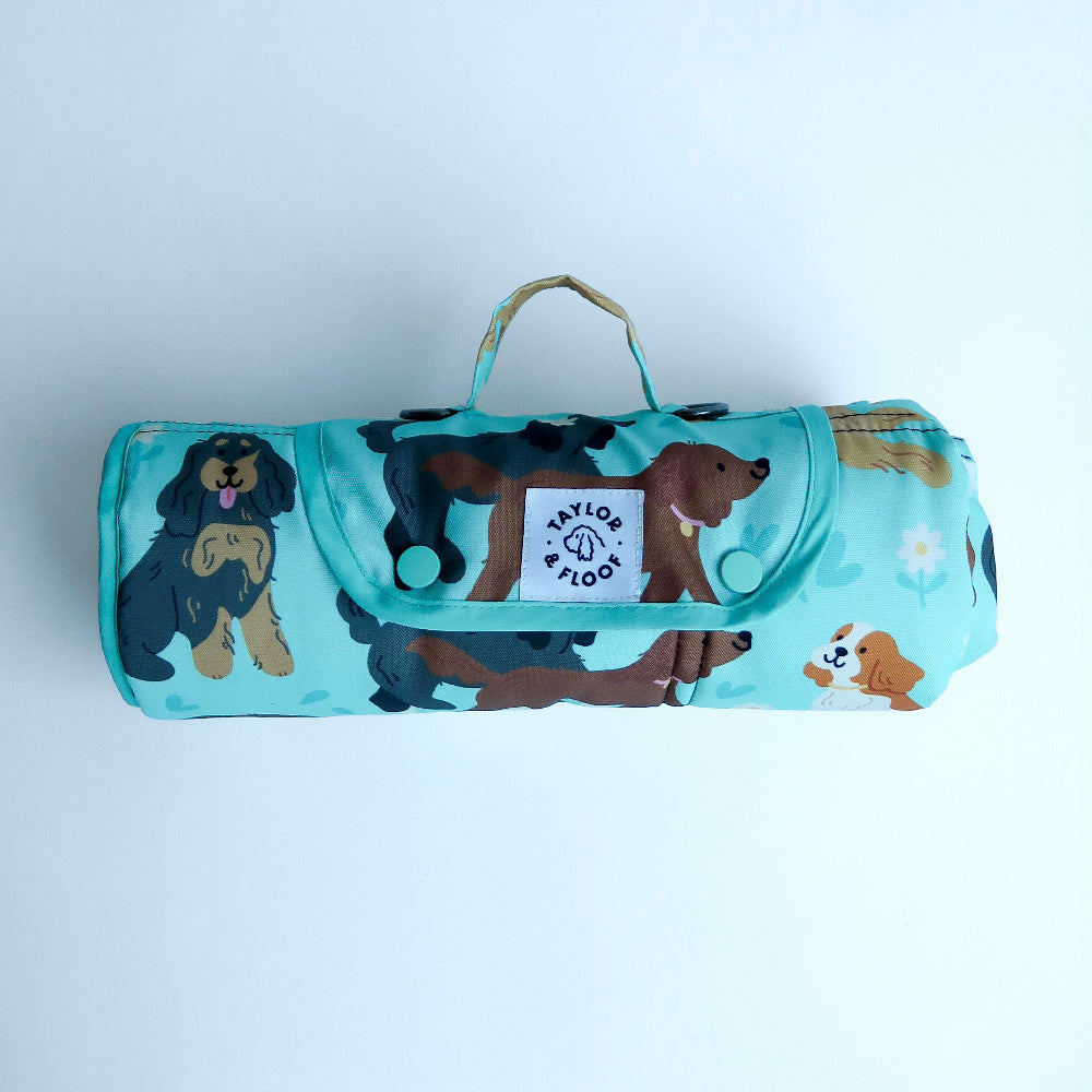 Roll-up blanket with spaniel pattern and visible brand logo on a light gray background. Light blue waterproof dog settle mat featuring cute spaniel illustrations perfect for cocker, springer, and cavalier spaniel lovers.